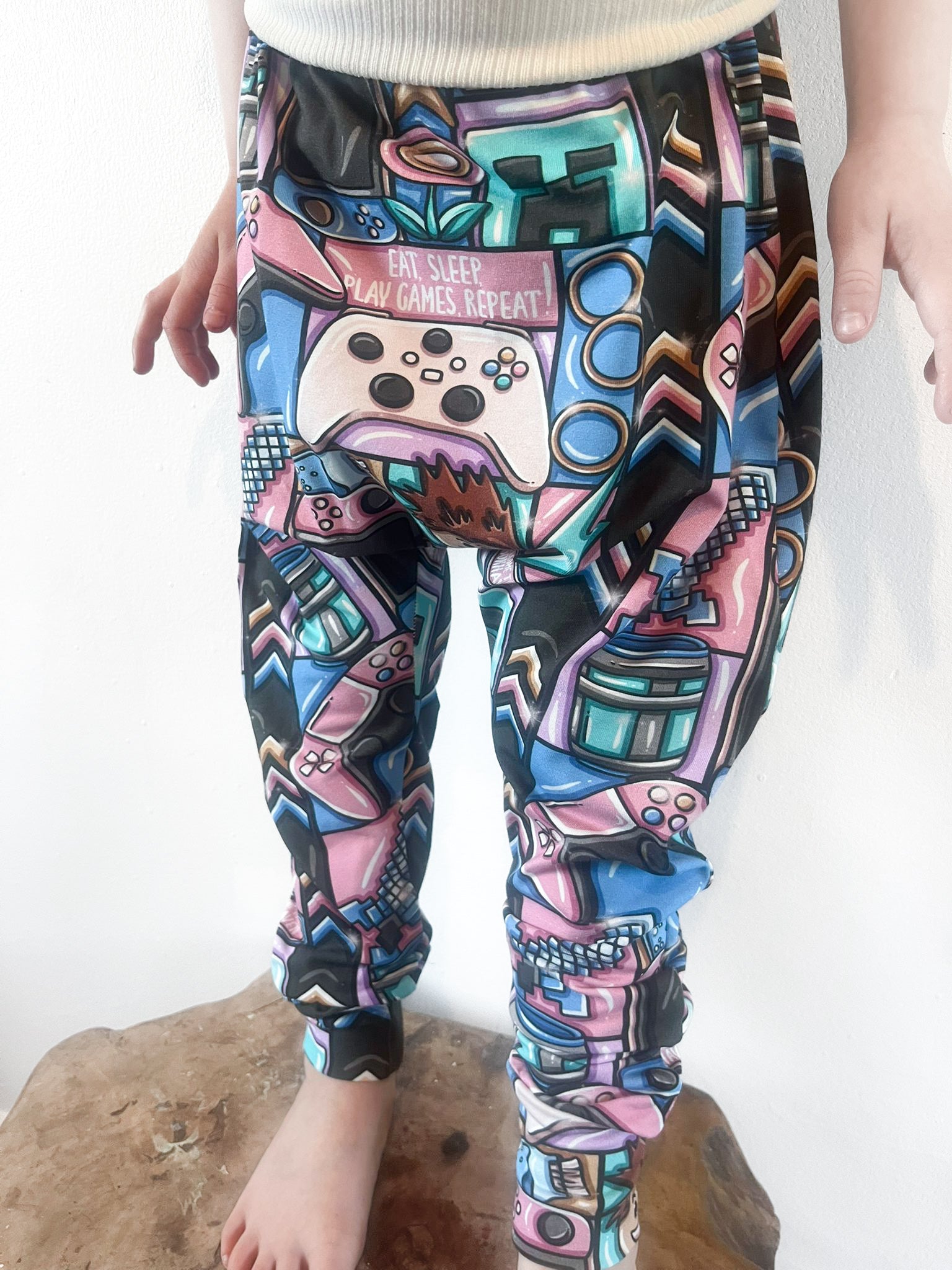 Child wearing baggy harem pants with colorful gaming-themed print featuring controllers, consoles, and gamer slogans