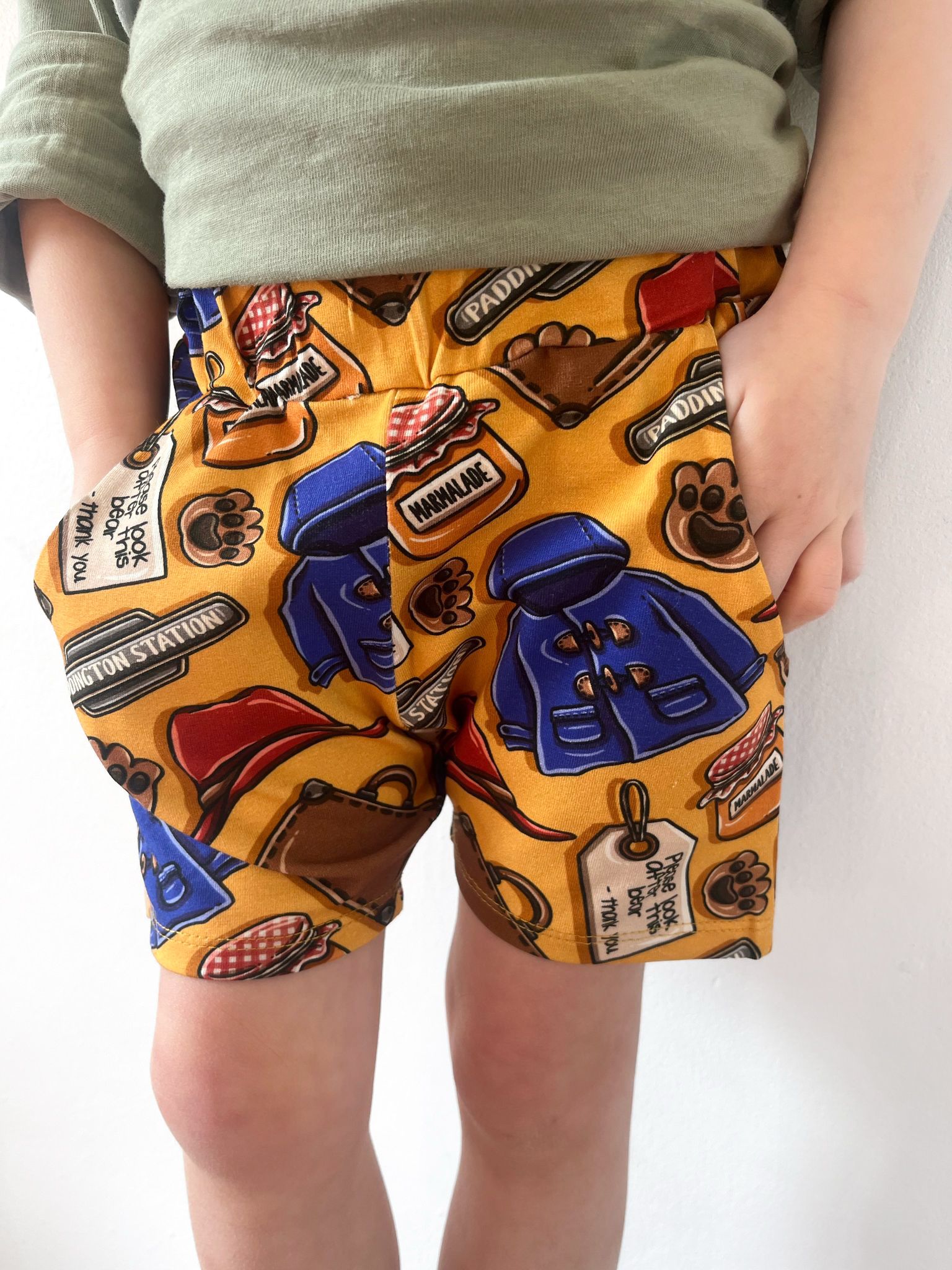 Child wearing British Bear -themed jogger shorts with golden-yellow background, blue coats, marmalade jars, and pockets.