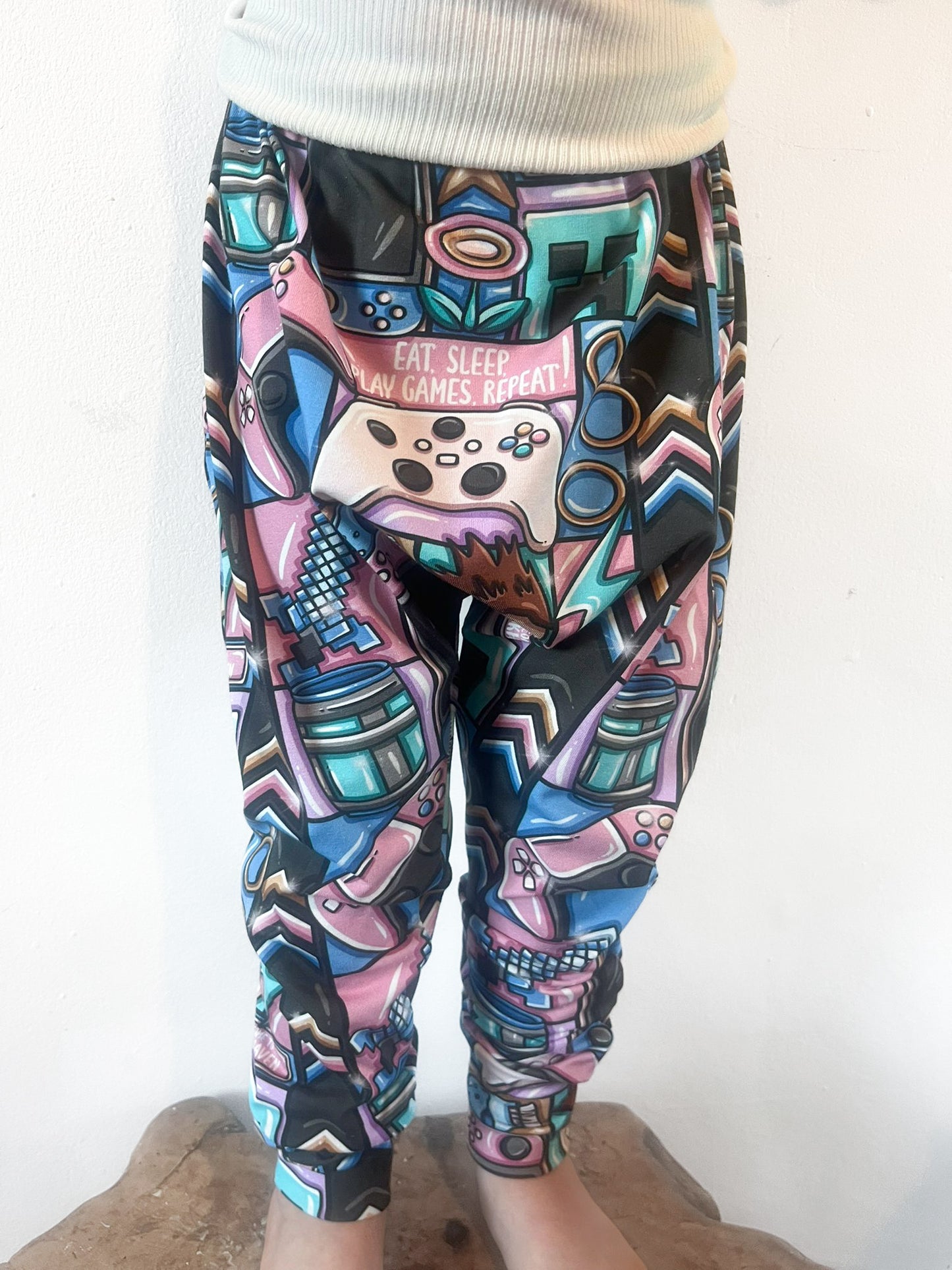 Child wearing baggy harem pants with colorful gaming-themed print featuring controllers, consoles, and gamer slogans