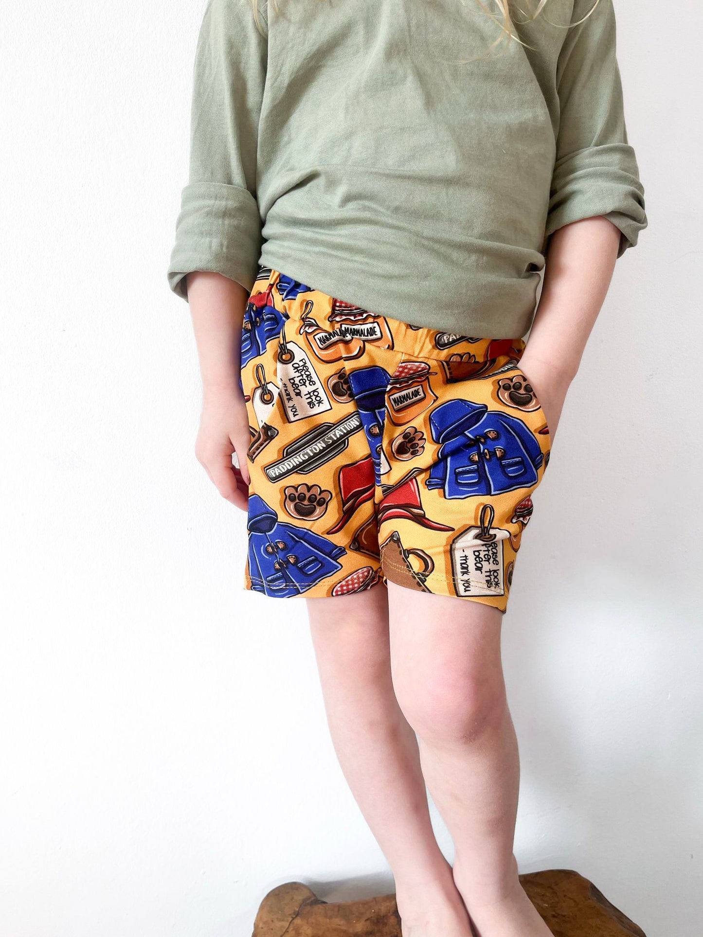 Child wearing British Bear -themed jogger shorts with golden-yellow background, blue coats, marmalade jars, and pockets.